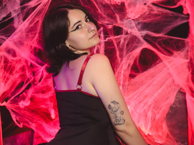MissTat with dark hair, nose ring, and black dress with red straps, showing tattoo on arm, surrounded by red, web-like, fabric. She looks over shoulder, sultry expression.