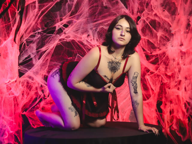 MissTat with dark bob, red straps, black dress, tattoos, kneeling on black circle, holding red strings, red and pink spider webs, looking at camera, showing tits and ass.