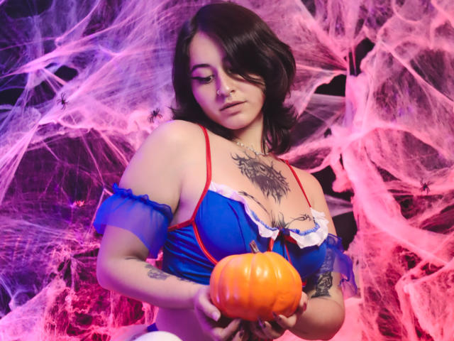 MissTat with dark hair, blue and red lingerie, holding a small orange pumpkin, tattoo on chest, tattoo on arm, looking down, surrounded by pink and purple spider webs.