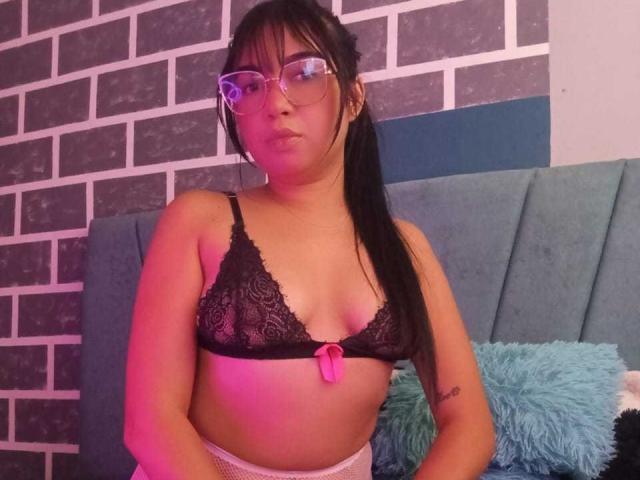 AliceSmity, a young woman with medium brown skin and black hair in a ponytail, wears clear glasses and a black lace bra with a small pink bow. She has a tattoo on her right forearm. Her bra reveals small, slightly sagging breasts. She stands in a room with a blue upholstered headboard, teal and blue pillows, and a brick-patterned wall. The lighting casts a pinkish-purple hue. Her expression is neutral, with slightly pursed lips.