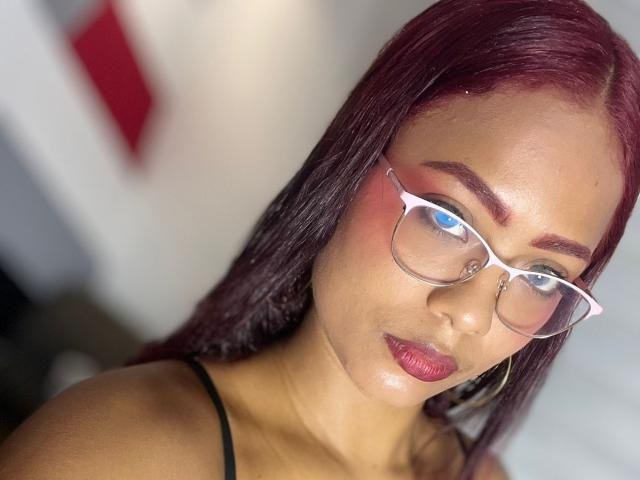 ChloeBrownOne with dark red hair, wearing glasses, has shiny red lipstick, pink eyeshadow, and is looking slightly to the side with a serious expression. She's wearing a black strap.