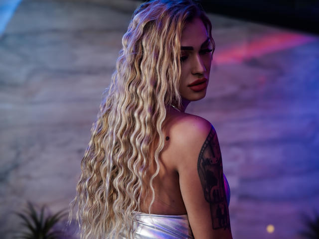 NicolSNaya with long, wavy blonde hair, looking over her shoulder, showing her bare back and tattooed arm, wearing a shiny silver dress, with her tits slightly visible.