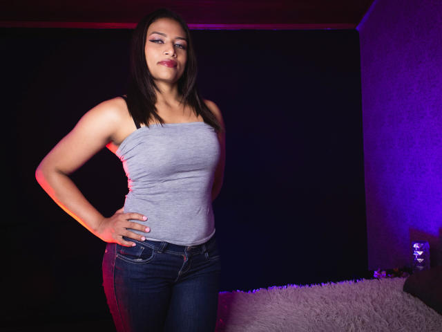 KellySmash with medium tits, gray tank top, and blue jeans, standing with hands on hips, dark hair, pink lipstick, purple and red lighting, looking confident, bedroom setting.