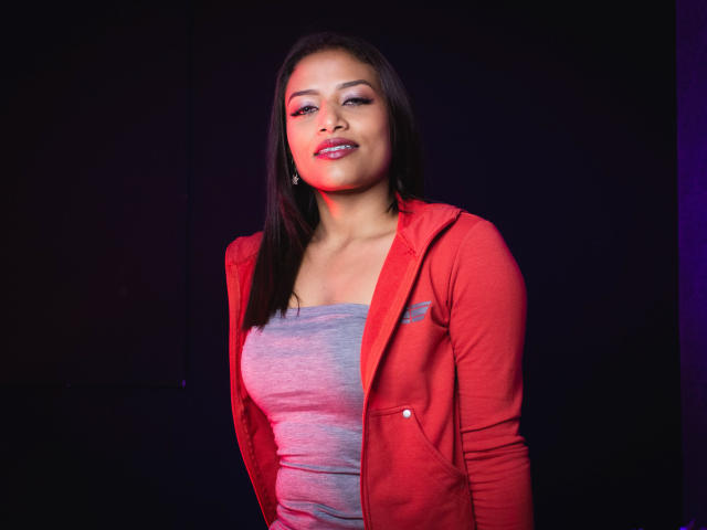 KellySmash with dark brown hair, red jacket, and gray top, smiling slightly, showing small tits, medium ass, and cute face, standing against a dark background.