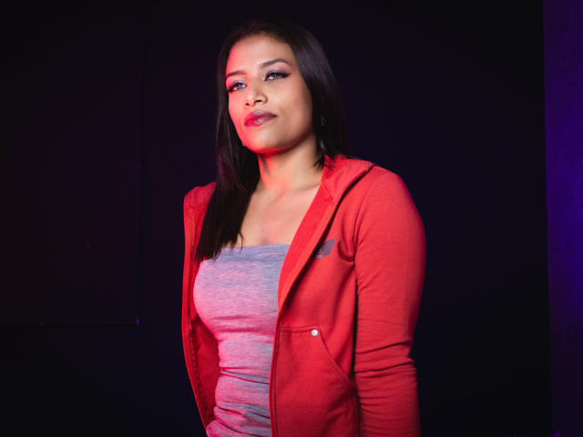 KellySmash with dark brown hair, wearing a red jacket over a gray tank top, has a confident, slightly smug look, with red lighting on her face and chest.