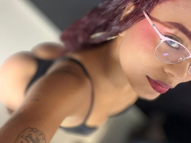 ChloeBrownOne with red hair, glasses, and dark lingerie, showing her ass and small tits, pink blush on cheeks, tattoo on arm, looking up at the camera.