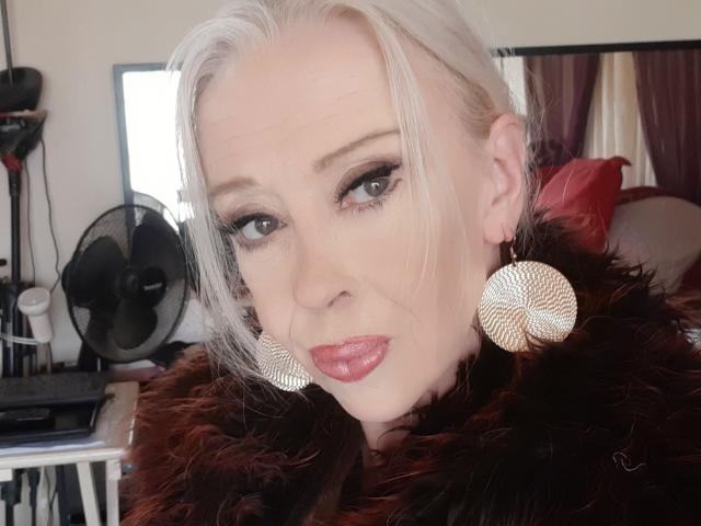 IssahneMay with pale skin, platinum blonde hair, and red lipstick, wearing large circular earrings and a brown fur collar, has a sexy, confident look. Her tits are not visible.