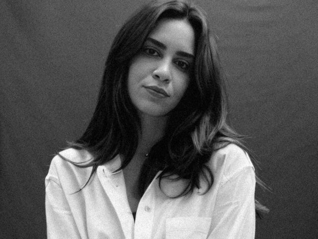 YourgirlGabriela with long, wavy dark hair, wearing an open white shirt, showing a slight smile, and soft eyes in a black-and-white photograph. Her tits are slightly visible.