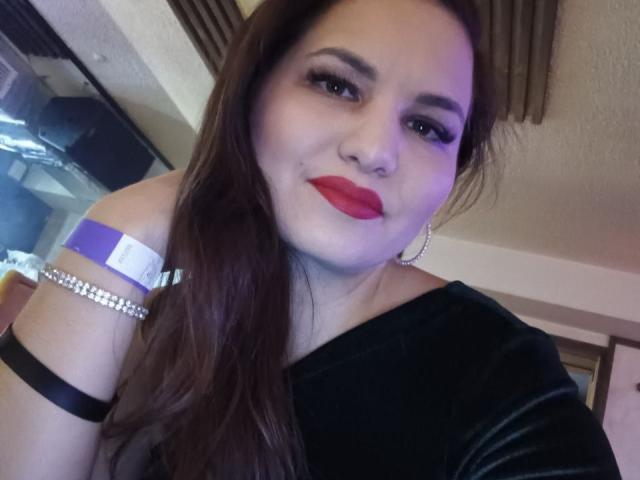 AlinasissyX with red lipstick, long brown hair, black off-shoulder top, silver arm bracelet, hoop earrings, smiling at camera, purple light on face, brown curtain in background.