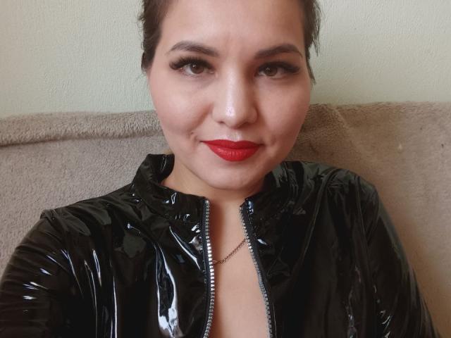 AlinasissyX with red lipstick, black shiny latex jacket, and visible cleavage, smiling slightly, brown eyes, and dark hair in a bun, wearing a thin necklace.