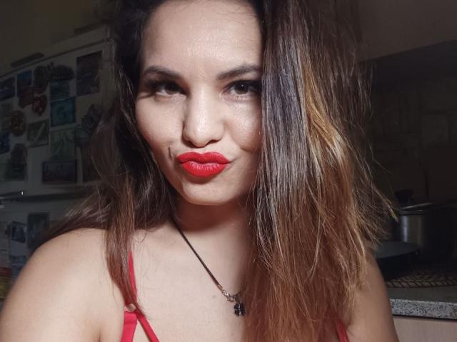 AlinasissyX with red lipstick, slightly wavy brown hair, and a black necklace, wearing a red top, has a smirking expression in a kitchen.