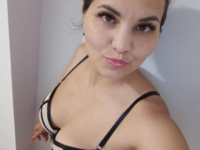 AlinasissyX with medium tits in a black and beige bra, pouty pink lips, dark eyebrows, and red nails, looking up at the camera with a slight smile.