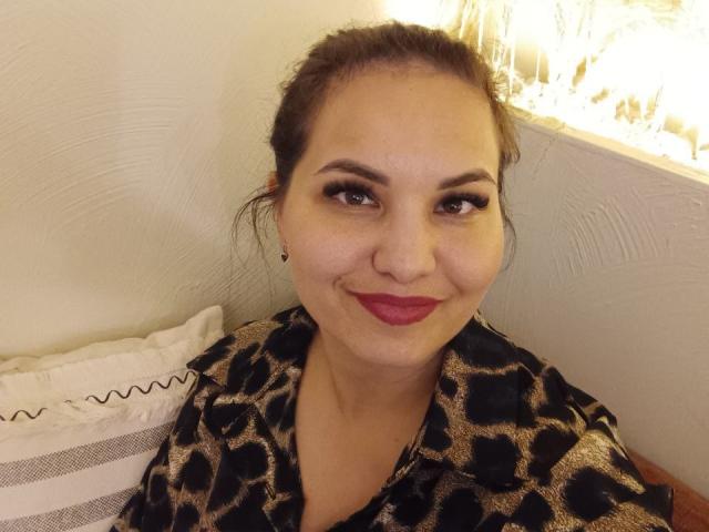 AlinasissyX has dark brown hair in a messy bun, wearing a leopard print shirt, with red lipstick and black eyeliner, smiling slightly. She has small tits and a cute face.