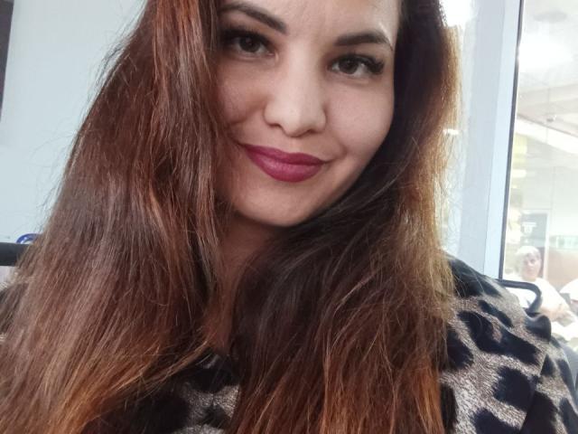 AlinasissyX with long brown hair, wearing a leopard print shirt, has red lipstick and a slight smile, looking directly at the camera. Her nose is small and her skin is light.