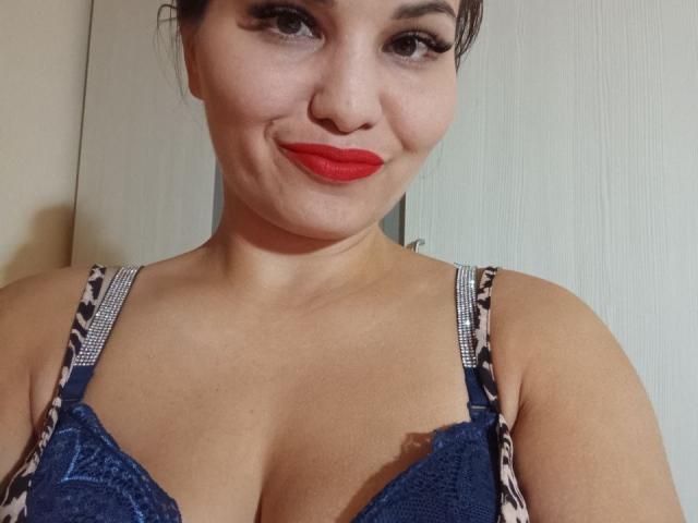 AlinasissyX with red lipstick, blue lace bra, and leopard-print straps, smiling at the camera, showing her cleavage, and brown eyes. She's got a cute, curvy face.