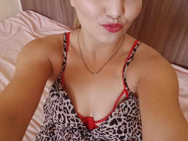 AlinasissyX with pouty red lips, wearing a leopard print dress with red trim, showing her tits in a low-cut top, necklace, and a red bra underneath.