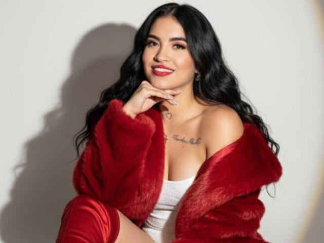 DaniellaPicc with long black hair, red fur jacket, white tube top, and red boots, smiling, hands near face, small heart tattoo on chest, medium tits.