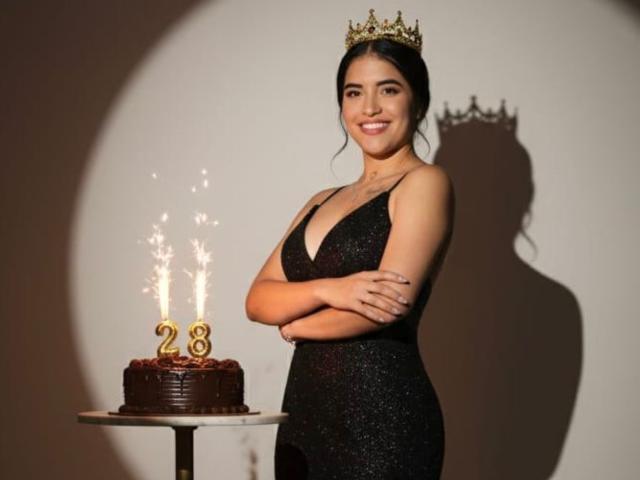 DaniellaPicc indossa un abito nero scintillante con una corona dorata, è in piedi con le braccia conserte, sorridendo a una torta al cioccolato con le candeline su un tavolo. Il seno è visibile..