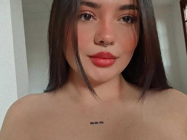 ValeriJoness has dark brown hair, a nose ring, and red lipstick. She's looking at the camera with a slight smile, showing a small black tattoo below her tits.