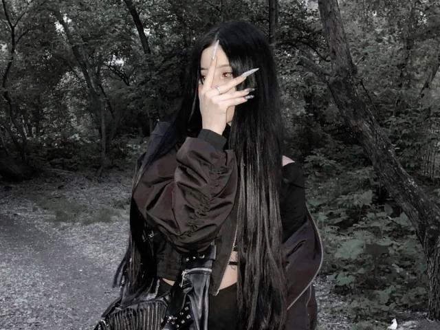 LollyCat with long black hair, wearing a black jacket and pants, covering her face with one hand, has long nails, standing in a forest with a gravel path.