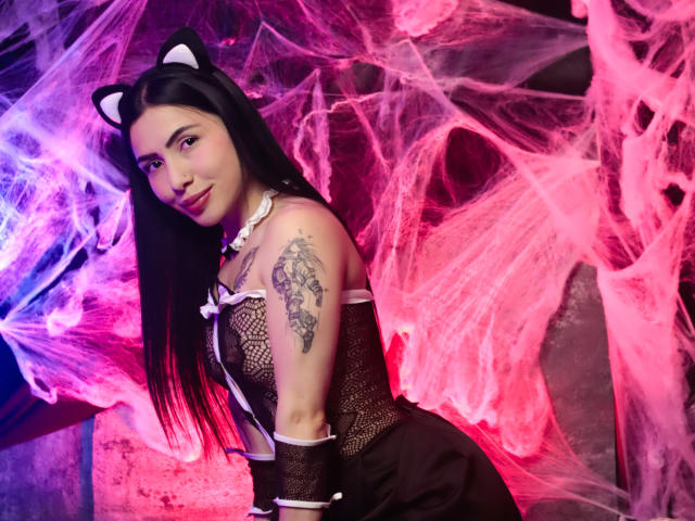 LiaVega with black hair, cat ears, fishnet top, white choker, tattoo on left arm, smiling, pink and purple smoke, black dress, looking over shoulder, sexy.