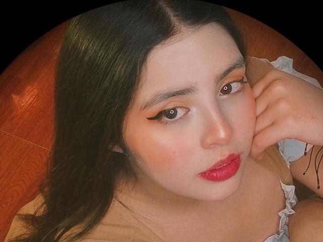 Paola Lyon ha la pelle chiara, occhi verde scuro con eyeliner nero, rossetto rosso, ed è sdraiata su una superficie arancione con una mano vicino al viso, guardando la fotocamera..