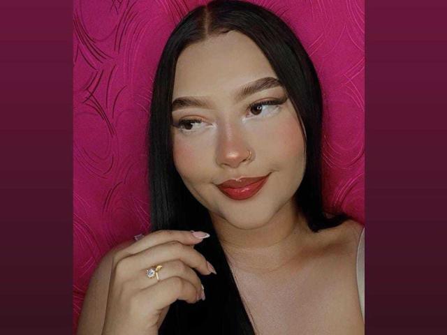 CandasLeroy with long black hair, brown eyes, red lipstick, and a nose ring, smiling slightly, holding a ring on her manicured nails, wearing a white top.