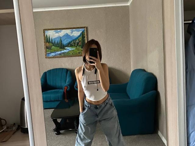 AmiaCrawford with small tits, wearing a white tank top and loose gray pants, taking a mirror selfie in a room with blue couches.