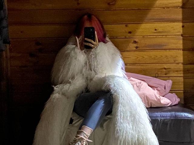AmiaCrawford with red hair, wearing a fluffy white coat, taking a selfie with a phone, in dark jeans and white lace-up shoes, sitting against a wooden wall.
