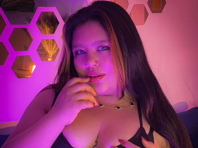 CaroHotcg with long brown hair, pink and purple lighting, black bra, nose ring, and gold necklace, biting finger, showing her tits, and looking at camera.