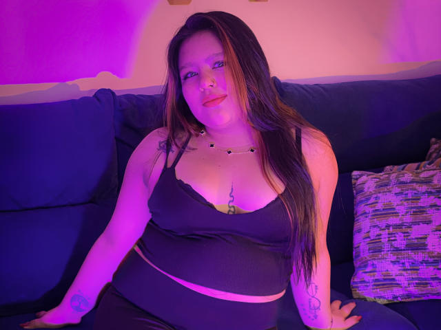 CaroHotcg with long brown hair, black top, and black pants, sitting on a blue couch, purple and pink lighting, smiling, visible tattoo on right arm, medium tits.