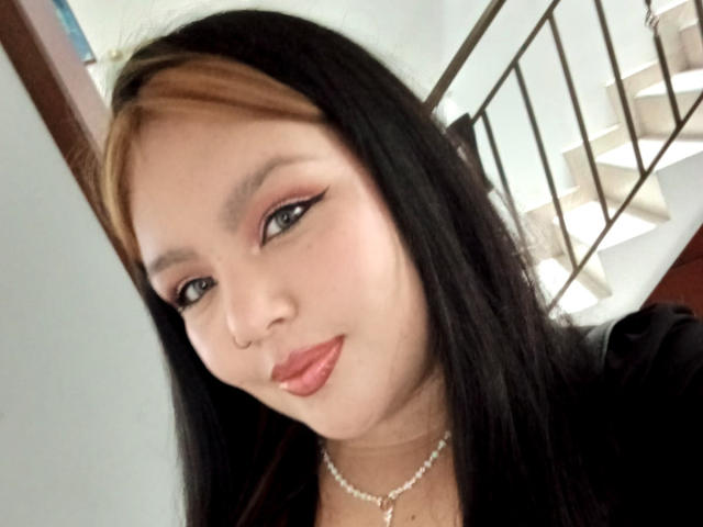CaroHotcg has black hair with a brown streak, pink eyeliner, and glossy red lips. She wears a pearl necklace and smiles slightly. Her nose is pierced.