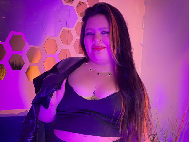 CaroHotcg with long brown hair, purple and pink lighting, black strapless top, black leather jacket, necklace, smiling, showing her tits, holding jacket, pink lipstick, tattoo under top.