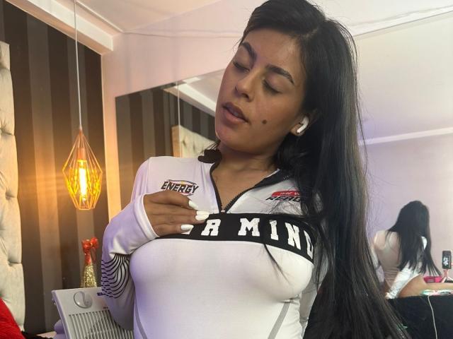 CamilePretty with long black hair, wearing a white sports shirt, covering her tits with one hand, white earbuds in, looking down, in a softly lit room, showing a beauty mark.