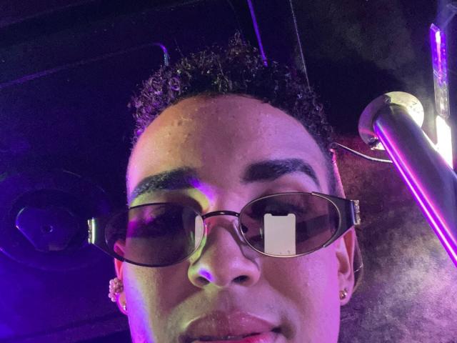 Marcuszion, avec ses cheveux noirs bouclés, ses lunettes et ses petits boucles d'oreilles, arbore un léger sourire, avec un éclairage violet et bleu sur son visage. Son nez est bien centré dans le cadre..