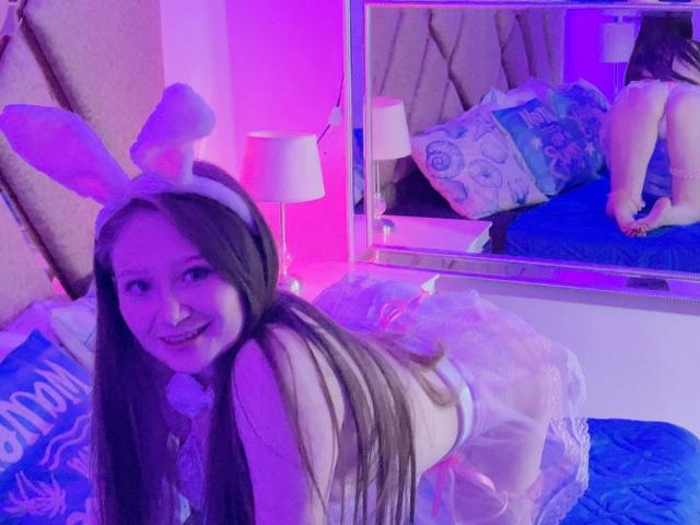 CuteIrinaX with long brown hair, smiling, wearing white bunny ears, sheer pink dress, and white bow, kneeling on bed, purple and pink lighting, reflection in mirror showing her ass.