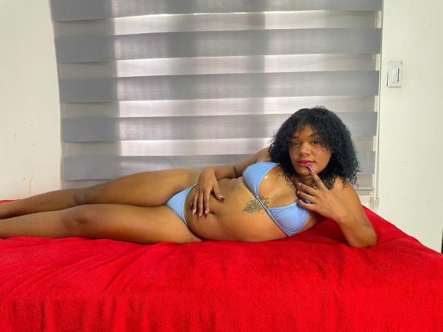 BigAssXx69 lying on a red bed, wearing a light blue bikini, showing her tits and ass, with curly black hair, a tattoo on her right side, and red lipstick.
