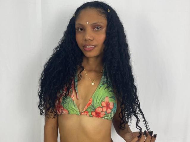 BigAssXx69 with long curly black hair, wearing a floral bikini top, small brown eyes, dark skin, gold necklace, and a small dot on her forehead, smiling.