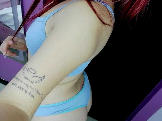 SamaraCuttie with red hair wears a light blue bra and matching panties. Her right arm, showing a tattoo of a winged heart with cursive text, is slightly raised. Her skin is pale, and the tattoo is black. The photo focuses on her upper body and right arm. The background is purple and black. Her hair is straight and red, visible over her shoulder. The image is a close-up, slightly angled.