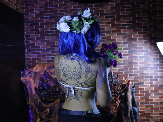 KoralineMiller with blue hair, white flower crown, holding purple roses, back tattoo of intricate black design, wearing black belt, standing in front of brick wall.