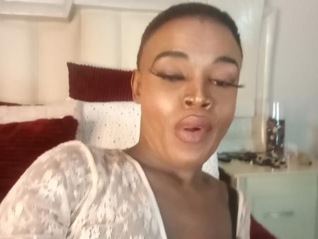 LucileCarrie has dark skin, full lips puckered, and is wearing a white lace top. She has eyeliner, short hair, and is making a kissing face. Her breasts are visible.