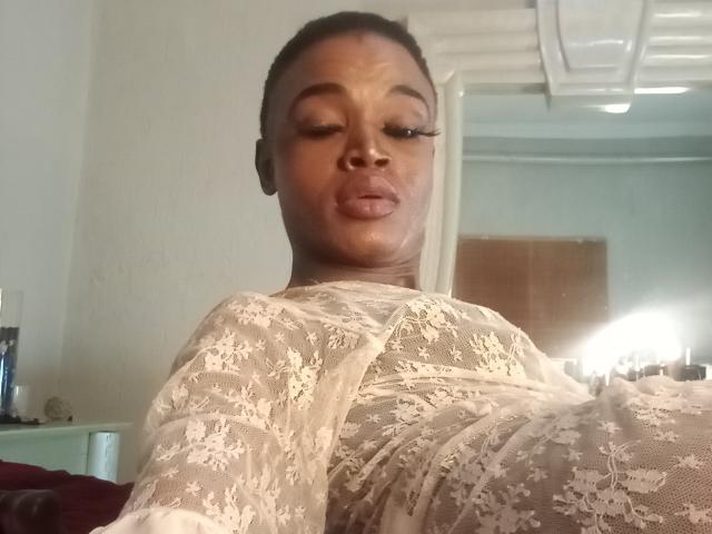 LucileCarrie, dark-skinned woman, pouty lips, wearing white lace dress, visible breasts, black hair in bun, eyeliner, light in background, holding camera, selfie, bedroom setting.