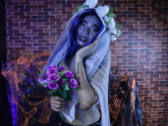 KoralineMiller with blue face paint, white flower headpiece, and veil, holding purple roses, has tattoo on left hand, wearing sheer top, against brick wall, looking surprised.