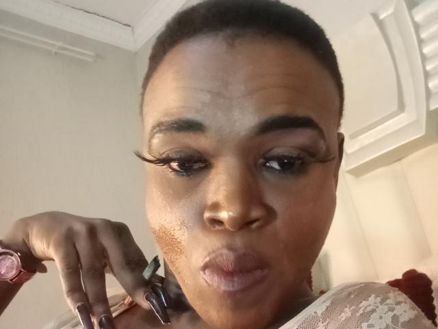 LucileCarrie, a dark-skinned woman with short hair, puffy lips, and thick black eyelashes, has a brownish acne mark on her right cheek, wearing a lace top.