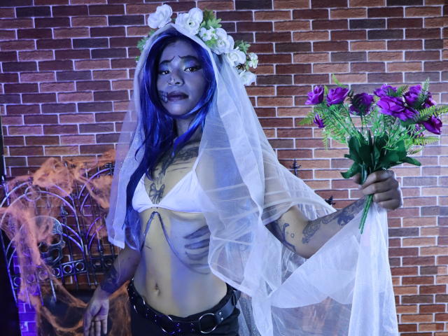 KoralineMiller with blue hair, gray face paint, white flower crown, sheer veil, white bra, black belt, holding purple roses, tattoos on arms, brick wall background, smoke.