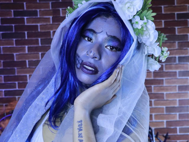 KoralineMiller with blue face paint, purple lipstick, and white flower headpiece, wearing a translucent veil, touching her face, showing tattoo on her hand. Brick background.