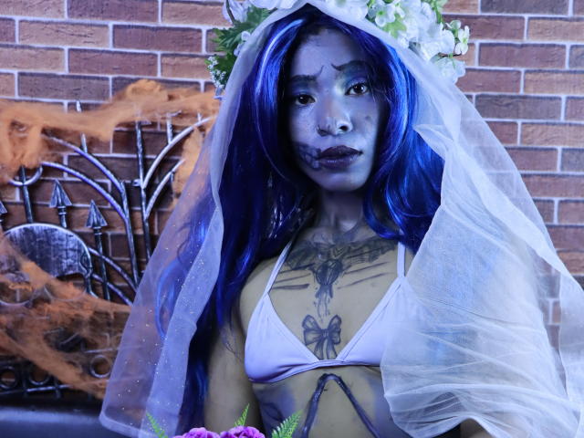 KoralineMiller with blue-painted face, blue hair, white veil, flower crown, white bikini top, tattoos, holding flowers, standing in front of brick wall, metal fire sculpture.