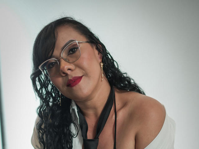 Mulher latina com cabelos pretos cacheados, batom vermelho, óculos, brincos de ouro, blusa branca com um ombro descoberto, gravata preta, sorrindo para a câmera, mostrando seus ombros e colo..