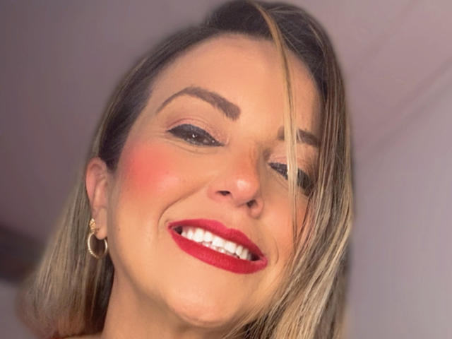LafolieDouce69 has blonde hair, red lipstick, and a smile showing white teeth. She's winking with a single eyebrow raised, wearing gold hoop earrings, and has a slight blush.