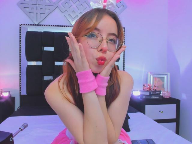 KattyHards with puffy pink lips, glasses, pink wristbands, and pink pigtails, pouting, hands on cheeks, in a bedroom with purple and pink lights.